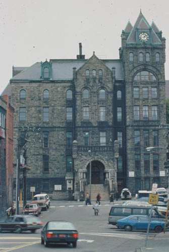 Front facade of St. John's Court House showing the three-storey, Duckworth Street facade, characterized by a central projecting bay with arched entrance, a series of blind arches, a square tower at one corner, and a massive round tower at the other. © Parks Canada | Parcs Canada