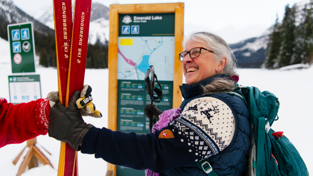 Une personne sourit à quelqu’un qui lui tend ses skis alors qu’elle se tient devant un panneau directionnel au lac Emerald.