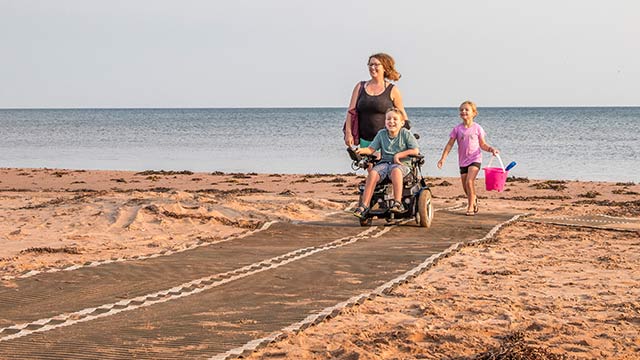 A child in a wheelchair, another child holding beach toys, and an adult, are returning from the beach using a universal beach mat