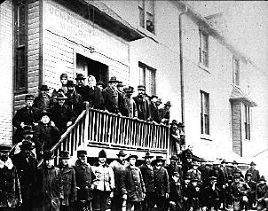 A black-and-white photo of people standing on the street and the staircase in front of a building with Dominion Government Immigration Hall painted above the door.