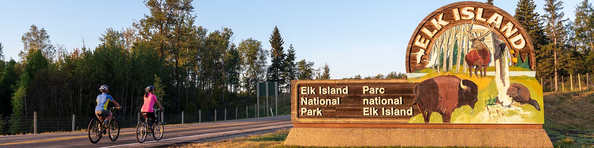 Road cyclists bike past the entrance sign at the north gate of Elk Island National Park. Elk Island National Park