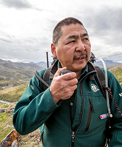 Parks Canada employee talking on a two-way radio, mountains and fields in the background.