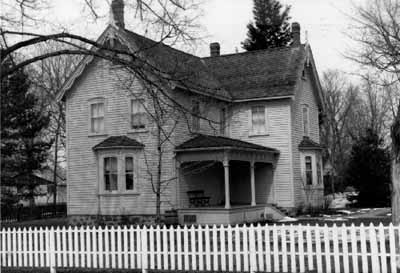 Vue de la façade sud de la maison Bethune, qui montre le bardage à clins des murs extérieurs, l’arrangement symétrique des fenêtres, les baies vitrées et la véranda, 1989. © Agence Parcs Canada / Parks Canada Agency, 1989.