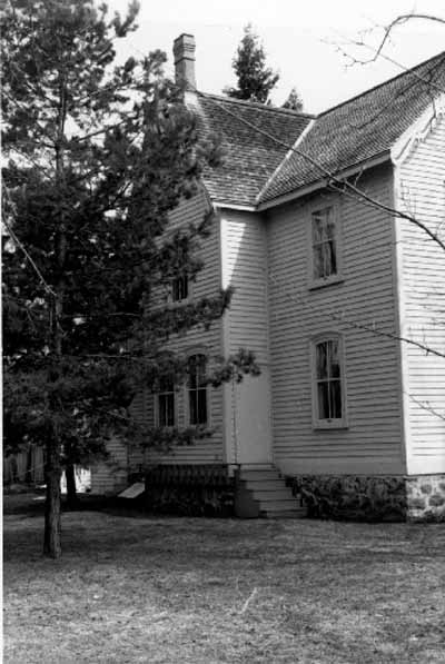 Vue de la façade ouest de la maison Bethune, qui montre le toit à deux versants très incliné, 1989. © Agence Parcs Canada / Parks Canada Agency, 1989.