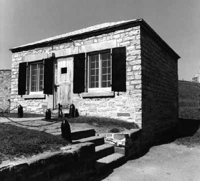 Vue de la façade générale du bâtiment 16, qui montre la volumétrie rectangulaire de plain-pied, de petite taille, coiffée d’un toit en croupe bas, 1980. (© Parks Canada Agency / Agence Parcs Canada, 1980.)