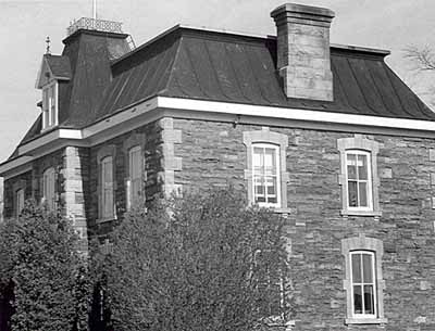 Corner view of the Office Building, showing the sandstone exterior walls laid in random courses and the mansard roof with chimneys, 1985. © Parks Canada Agency / Agence Parcs Canada, 1985.