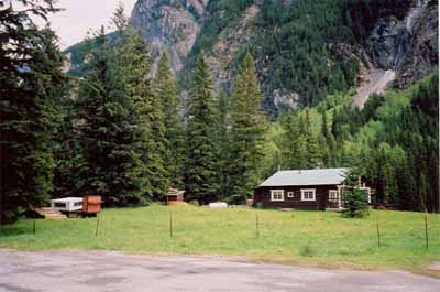 Panoramic view of Ranch-cabin and surroundings from the south driveway demonstrating its scenic location in the Yoho Valley, adjacent to flat pasture land on one side and a tall stand of coniferous trees on the other, 1999. © Cultural Resource Services, Calgary/Ressources culturelles, Calgary, 1999.