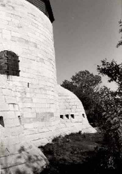 Side view of Cathcart Tower, showing the massively constructed, smooth, steep exterior walls built of tightly fitting limestone blocks and two of the four evenly spaced caponiers around the base, 1992. © Parks Canada Agency / Agence Parcs Canada, 1992.