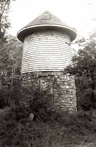 General view of the Water Tower, showing its tapered cylindrical shape, low-pitched conical roof, cedar-shingle cladding and rubble-stone base, 1992. (© Parks Canada Agency / Agence Parcs Canada / Historica Resources Ltd., 1992.)