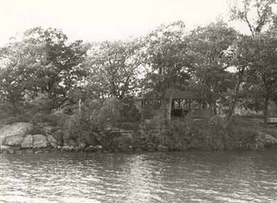 View of the Adelaide Island Picnic Shelter from the river, showing its relatively isolated island location on a heavily treed site, 1992. © Parks Canada Agency / Agence Parcs Canada / Historica Resources Ltd., 1992.