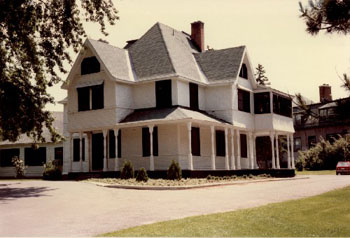 View of the main façade of Heritage House (Building 54), showing the large scale and massing of the house, which consists of a two-and-a-half storey structure with a verandah around two sides and a hip and gable roof, 1982. (© Agence Parcs Canada / Parks Canada Agency, Janet Wright and Monique Trepanier, 1982.)