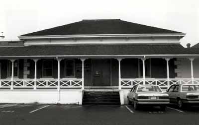 Façade of Building 37, showing its hipped roof, covered verandah, bracketed cornice and the segmentally-arched door and window openings with stone surrounds, 1989. © Ian Doull, Parks Canada Agency / Agence Parcs Canada, 1989.