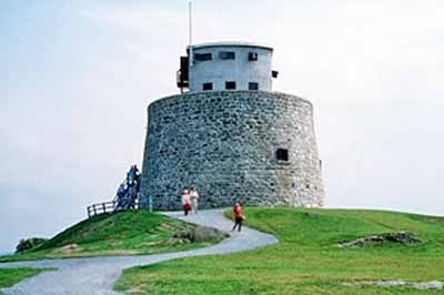General view of the Carleton Martello Tower showing the exterior walls built of rubble masonry which inclines slightly inwards as it rises to a parapet wall which is in turn topped by a two-storey concrete observation post, 1983. © Parks Canada Agency / Agence Parcs Canada, J. Steeves, 1983.