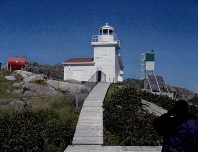 General view of West Point Light Tower, showing its colour pattern, as seen in the use of red shingles on the gable roof and white paint on its metal lantern and railing, 2006. (© Fisheries and Oceans / Pêches et Océans Canada, 2006.)