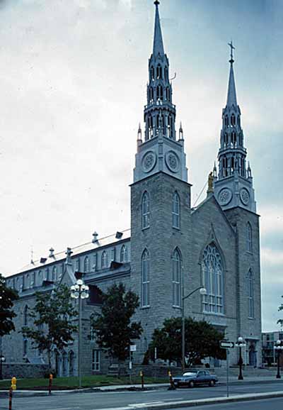 Vue générale de la basilique catholique Notre-Dame, qui montre la façade aux tours jumelles avec une grande fenêtre du côté ouest, 1989. (© Parks Canada Agency / Agence Parcs Canada, W. Duford, 1989.)