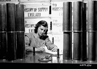 Woman surrounded by 25-pounder field gun cases writes on a notebook in a munitions factory (prob. Robert Mitchell Co.) © Nicholas Morant / National Film Board | Office national du film. Photothèque / Library and Archives Canada | Bibliothèque et Archives Canada