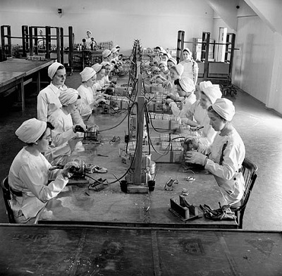 Women munitions workers solder fuse boxes at a long table in a munitions factory. © National Film Board of Canada | Office national du film du Canada. Photothèque / Library and Archives Canada | Bibliothèque et Archives Canada