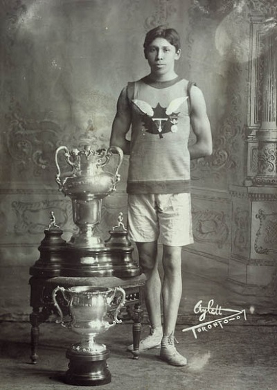 Tom Longboat, Canadian runner, standing beside trophies © Charles A. Aylett / Library and Archives Canada | Bibliothèque et Archives Canada / C-014090