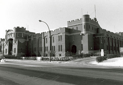 Corner view of the Mewata Armoury, showing one of the four large square, crenelated, three-storey, bastion like corner towers linked by continuous crenelated exterior walls, 1983. © Department of National Defence / Ministère de la Défense nationale, 1983.