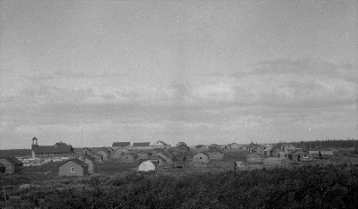 Historic photograph of Fort McPherson showing dwellings and the Anglican Church on the left of the image, standing still, ca. 1930 © J. F. Moran / Indian and Northern Affairs Canada | Affaires indiennes et du Nord Canada / Library and Archives Canada | Biliothèque et Archives Canada, a102490-v8