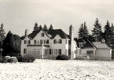 Vue du chalet du Premier ministre, qui montre ses cheminées en pierre et sa véranda ouverte, 1985. © Parcs Canada / Parks Canada, 1985.