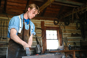 A Parks Canada employee in historic costume uses a hammer to strike a red-hot piece of metal on an anvil.