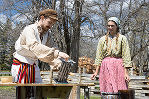 A Parks Canada employee in historic costume tilts a metal candle mould on a wooden table as another Parks Canada employee in historic costume watches.