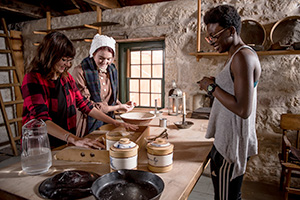 A visitor flattens dough on a wooden tray as a Parks Canada employee and visitor watch.