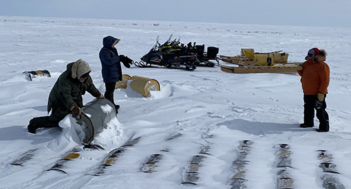One person rolls a large metal drum in the snow while two others stand. In the background, two snowmobiles with sleds behind them are seen.