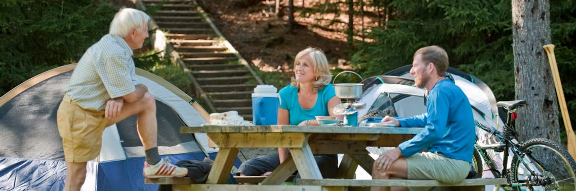 A group of three adults enjoy a meal at the picnic table at their campsite in Point Wolfe campground.