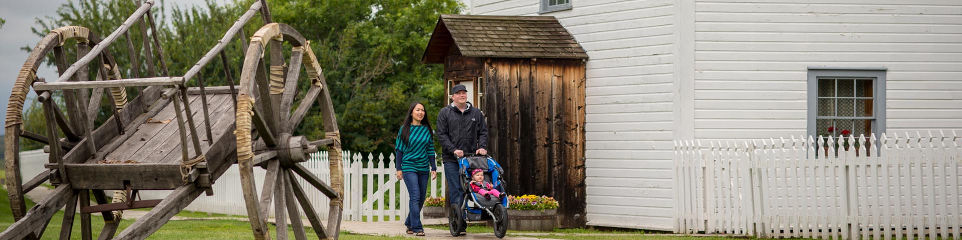 Two adults with a stroller walking along the boardwalk by the church and rectory at Batoche National Historic Site.