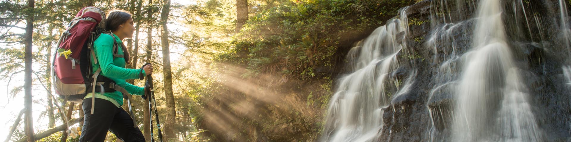 A female hiker marvels at the picturesque waterfall at Bonilla Creek, on the West Coast Trail, as the sun’s rays shine through the trees. Pacific Rim National Park Reserve.